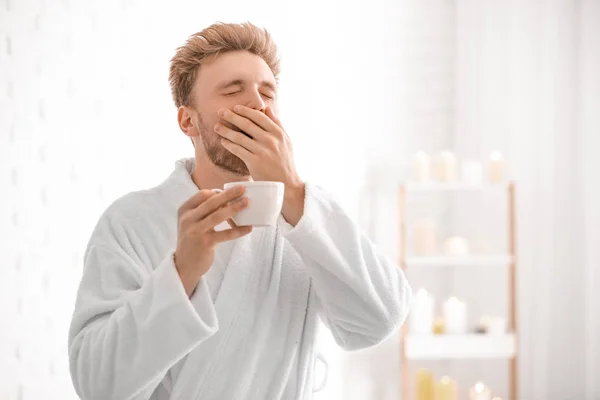 Sleepy young man in bathrobe drinking coffee at home