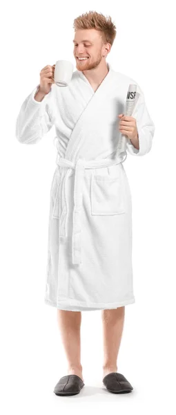 Handsome young man in bathrobe holding cup of coffee and newspaper on white background — Stock Photo, Image