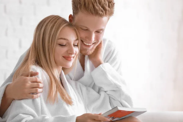Jovem casal feliz em roupões de banho leitura revista em casa — Fotografia de Stock