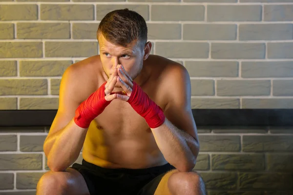 Strong male boxer sitting on bench against brick wall