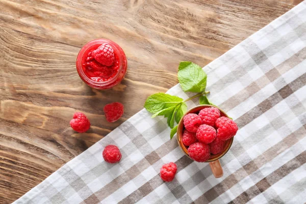 Cup with fresh raspberries and jam in jar on wooden table — Stock Photo, Image