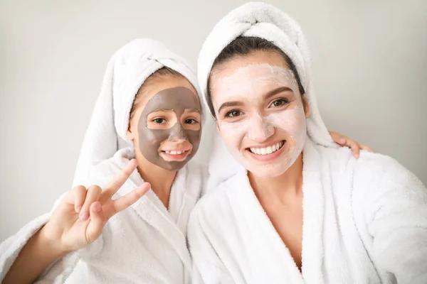 Mother and her little daughter in bathrobes and with facial masks against light background