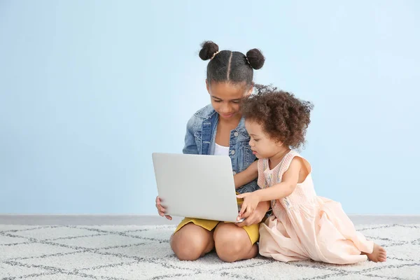 Cute African-American sisters with laptop watching cartoons near color wall — Stock Photo, Image