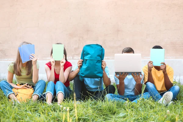 Mladí studenti skrývají obličeje při sezení na trávě venku — Stock fotografie