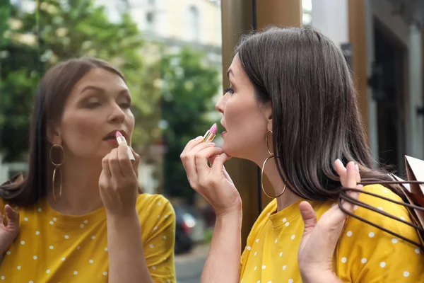 Beautiful woman applying lipstick while using shop-window as mirror — Stock Photo, Image