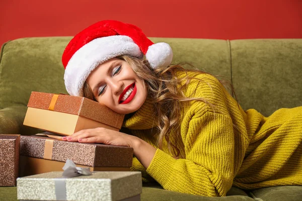 Beautiful young woman with Christmas gifts lying on sofa against color background — Stock Photo, Image
