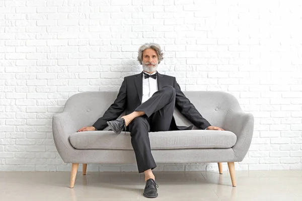 Модный пожилой человек, сидящий на диване у стены из белых брик — стоковое фото
