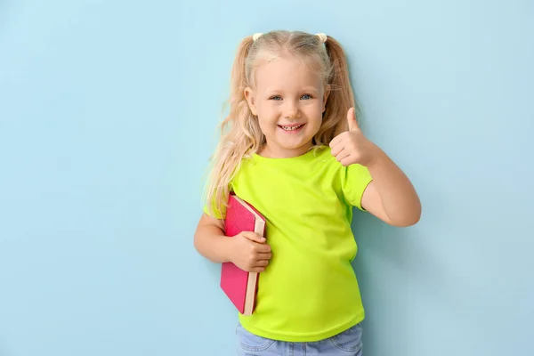 Χαριτωμένο μικρό κορίτσι με το βιβλίο δείχνοντας αντίχειρας-up στο χρώμα φόντο — Φωτογραφία Αρχείου