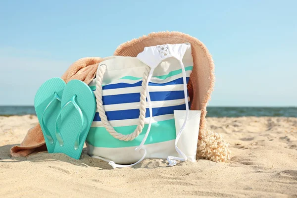 Bag with flip-flops, swimsuit, hat, towel and cosmetics on sand beach — Stock Photo, Image