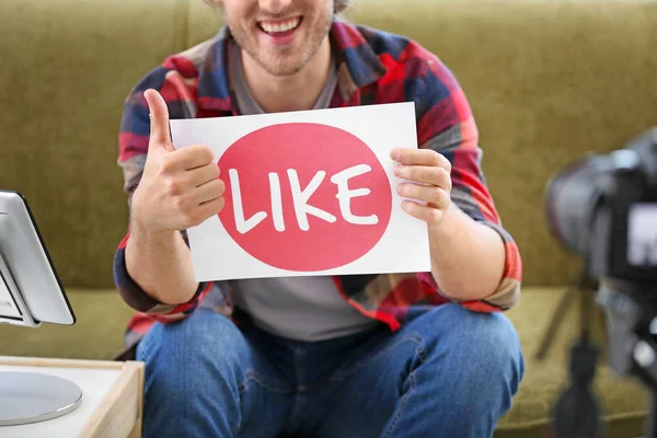 Male blogger holding paper with text LIKE while recording video at home — Stock Photo, Image