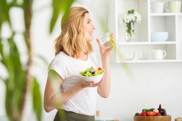 Mujer con ensalada de verduras saludables y vaso de agua en la cocina —  Fotos de Stock