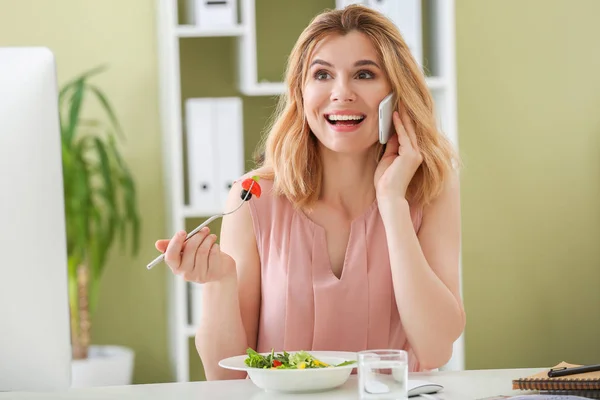 Woman eating healthy vegetable salad while talking by phone in office — Stock Photo, Image