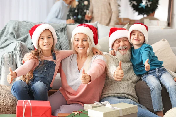 Abuelos felices y niños pequeños en sombreros de Papá Noel mostrando gesto de pulgar hacia arriba en casa — Foto de Stock