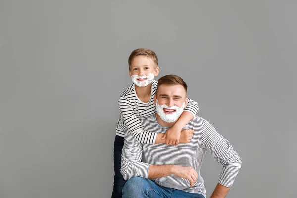 Father and his little son with shaving foam on faces against grey background — Stock Photo, Image