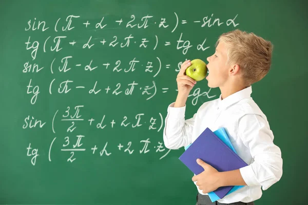 Niño comiendo manzana cerca de la pizarra escolar en el aula — Foto de Stock