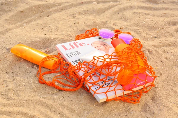 Сумка с кремом от загара, солнцезащитными очками и модным журналом на песчаном пляже — стоковое фото