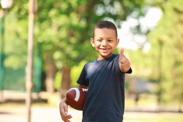 Cute little African-American boy with rugby ball showing thumb-up gesture in park — Stock Photo, Image