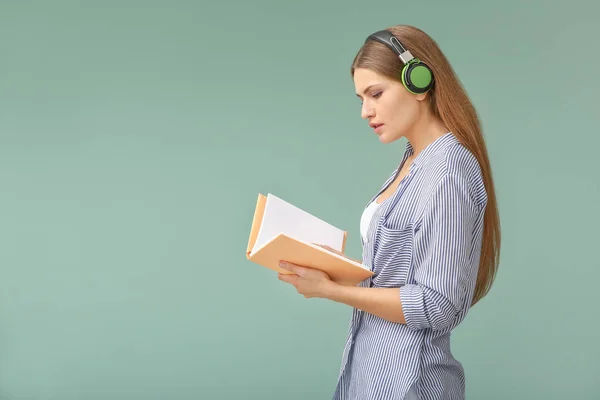 Young woman listening to music and reading book on color background — Stock Photo, Image