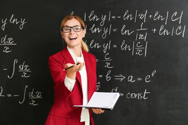 Schöne Mathelehrerin mit Buch neben Tafel im Klassenzimmer — Stockfoto