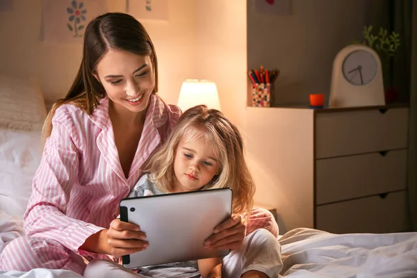 Happy woman and her little daughter with tablet computer on bed in evening — Stock Photo, Image