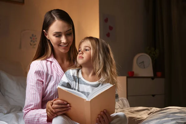 Happy woman with her little daughter reading book on bed in evening — Stock Photo, Image