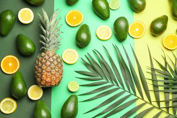 Fresh avocados, citrus fruits, pineapple and palm leaves on color background