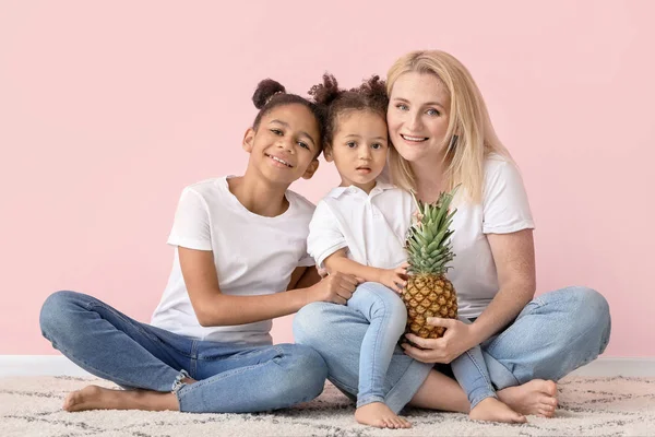 Happy woman and her little African-American daughters with pineapple sitting near color wall — Stock Photo, Image