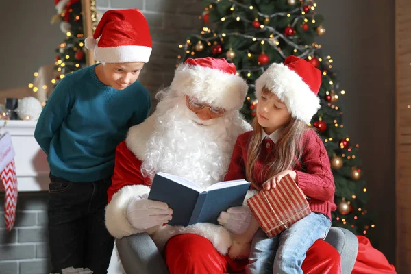 Santa Claus y los niños pequeños que leen el libro en la habitación decorada para Navidad — Foto de Stock