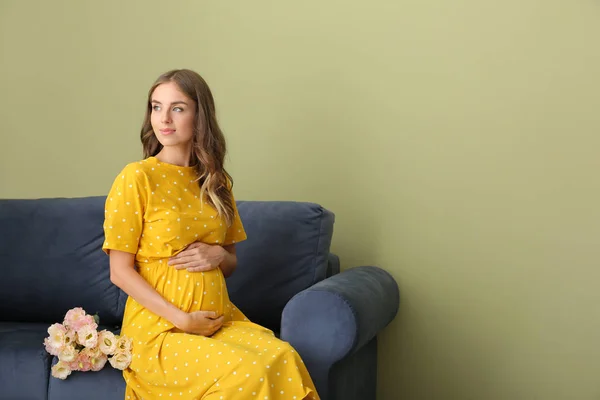 Mooie zwangere vrouw met boeket bloemen zittend op de Bank tegen kleur achtergrond — Stockfoto