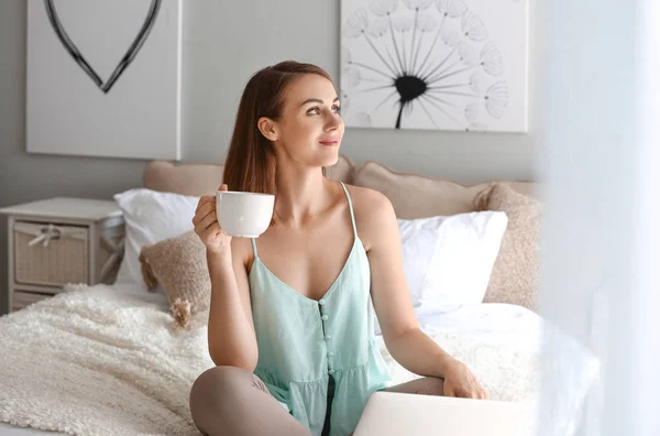 Beautiful young woman with laptop drinking tea at home — Stock Photo, Image