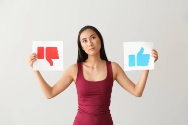 Düşünceli Asya blogger ışık arka plan üzerinde Like ve Dislike işaretleri ile kağıtları tutan