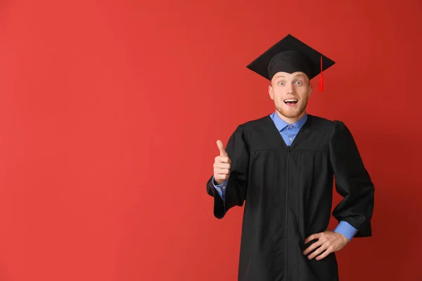 Young male student in bachelor robe showing thumb-up on color background