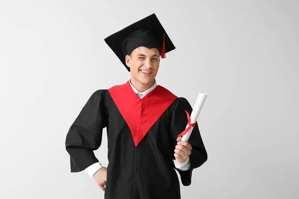 Young male student in bachelor robe and with diploma on light background — Stock Photo, Image