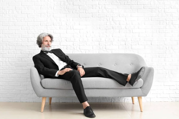 Uomo anziano alla moda seduto sul divano vicino al muro di mattoni bianchi — Foto Stock