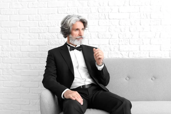 Fashionable senior man sitting on sofa near white brick wall — Stock Photo, Image