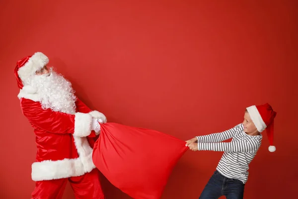 Kleiner Junge versucht, Tasche mit Geschenken von Weihnachtsmann vor farbigem Hintergrund zu nehmen — Stockfoto
