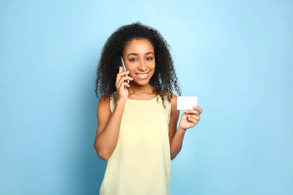 Portrait of beautiful African-American woman with business card talking by mobile phone on color background