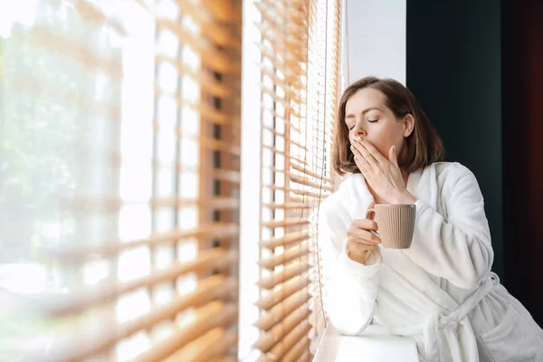 Sleepy young woman drinking coffee near window in morning — Stock Photo, Image