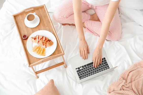 Beautiful young woman with laptop having breakfast in bed — Stock Photo, Image