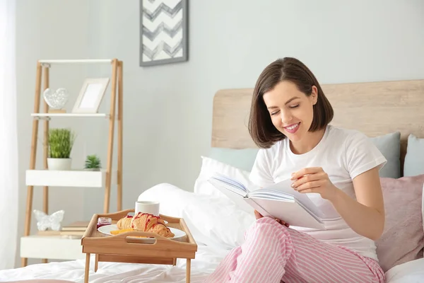 Morning of beautiful young woman reading book while having breakfast in bed — Stock Photo, Image