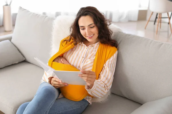 Young mother with little baby in sling reading book at home — Stock Photo, Image