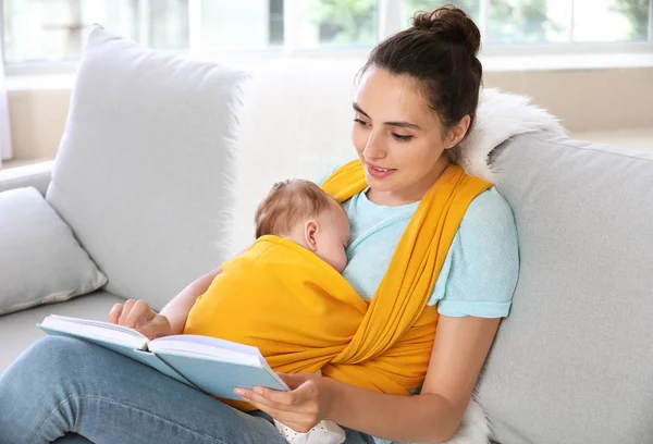 Young mother with little baby in sling reading book at home — Stock Photo, Image