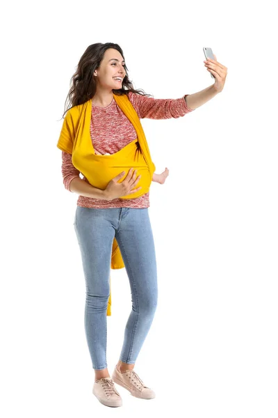 Young mother with little baby in sling taking selfie on white background — Stock Photo, Image