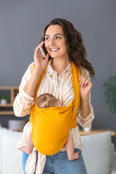 Young mother with little baby in sling talking by mobile phone at home — Stock Photo, Image