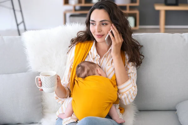 Young mother with little baby in sling talking by mobile phone at home — Stock Photo, Image