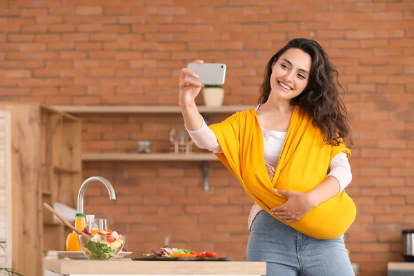 Jeune mère avec petit bébé en fronde prendre selfie dans la cuisine — Photo