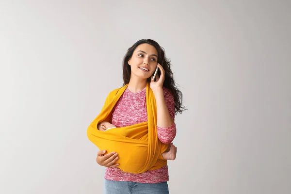 Young mother with little baby in sling talking by mobile phone on light background — Stock Photo, Image
