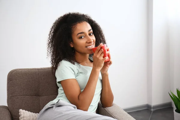 Beautiful African-American woman drinking tea in armchair at home — Stock Photo, Image