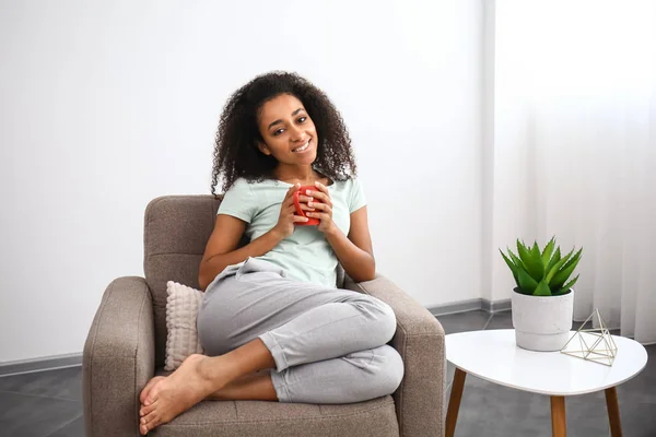 Beautiful African-American woman drinking tea in armchair at home — Stock Photo, Image
