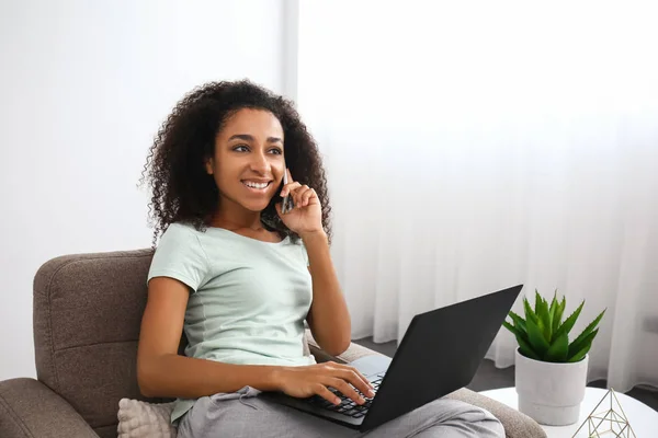 Beautiful African-American woman with laptop talking by mobile phone in armchair at home — Stock Photo, Image